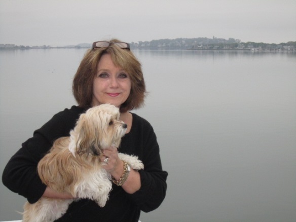 Leslie Luci at the Cape A woman with shoulder-length brown hair and glasses on her head smiles while holding a small tan and white dog. They stand outdoors in front of a calm, foggy lake with a distant shoreline visible under a gray sky. Community of Collectors