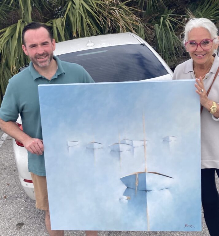 A man in a green polo shirt and a woman in glasses and a light top stand in front of a white car outdoors, smiling and holding a large, abstract painting featuring soft blue tones and sailboat shapes. Palm leaves are visible in the background.