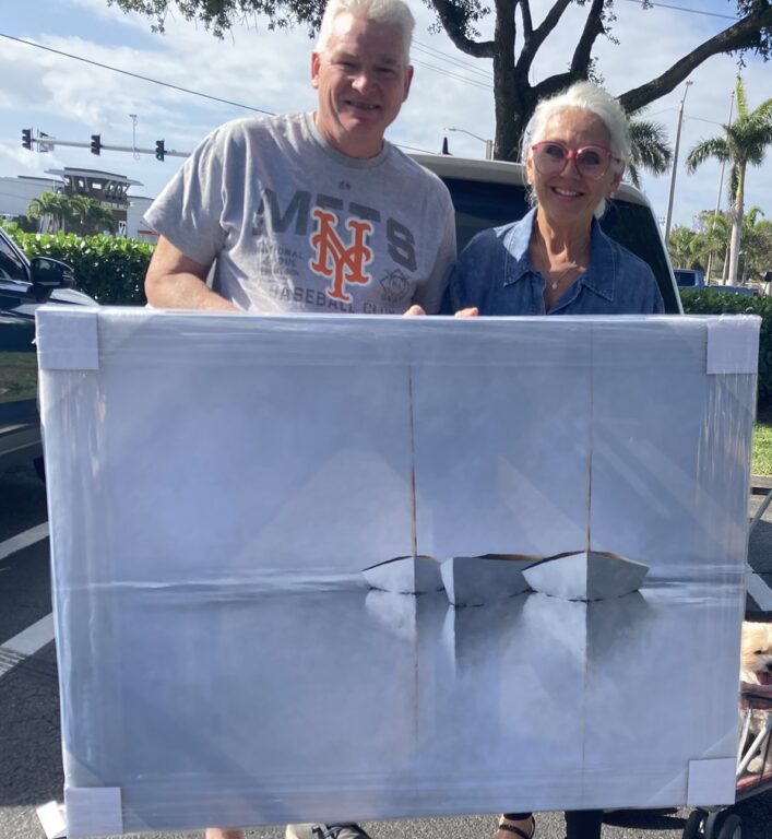 A smiling man in a gray New York Mets t-shirt and a woman in sunglasses and a blue shirt stand outdoors, holding a large, wrapped rectangular package with an abstract gray-white design. Palm trees and a parking lot are visible in the background.