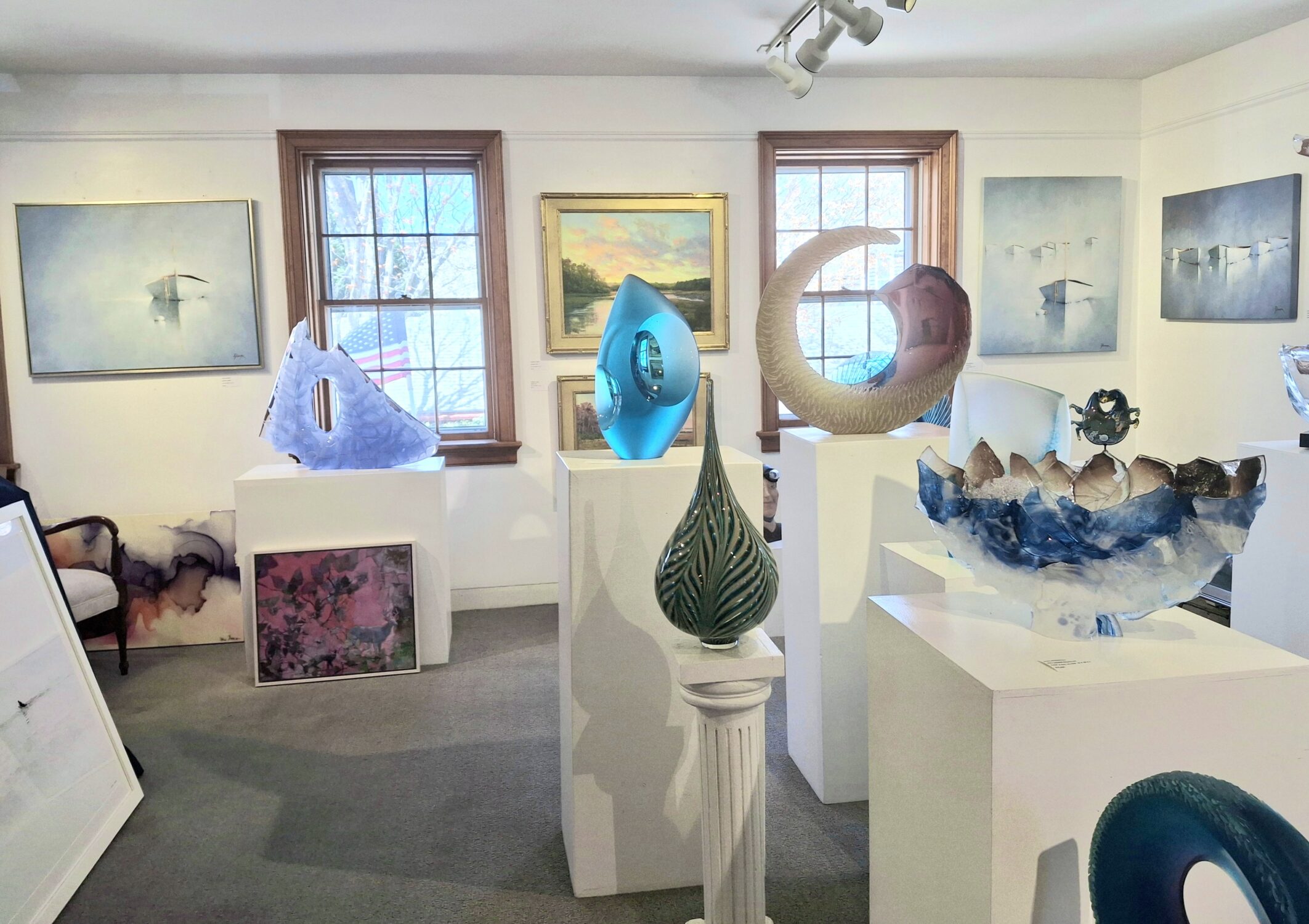 A bright art gallery room is one of the most inspiring locations, displaying abstract glass sculptures on white pedestals and soft-colored landscape paintings. Sunlight streams through two windows, illuminating the varied textures and forms of the modern artwork.