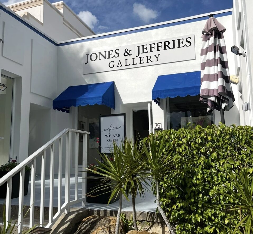 The exterior of the Jones & Jeffries Gallery, one of the area's standout locations, features white walls, blue awnings, and a striped umbrella. A sign in front reads “We are open.” Lush green plants border the walkway under a partly cloudy sky.