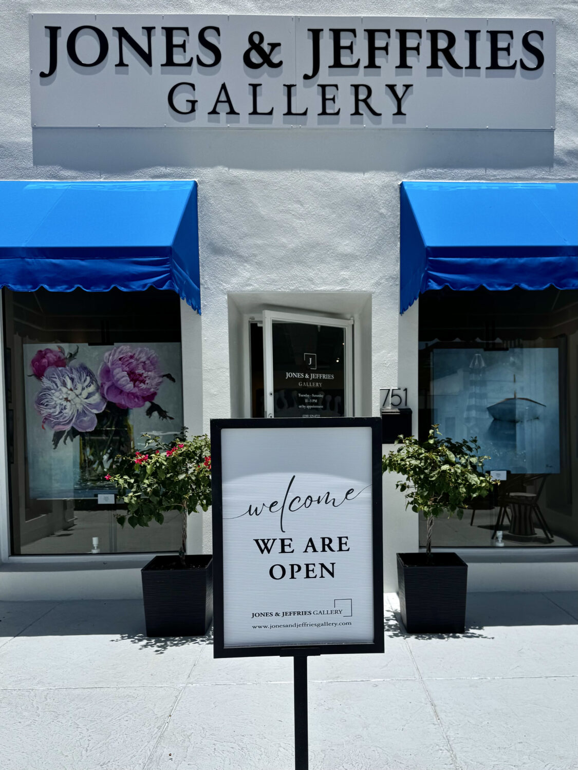Street view of Jones & Jeffries Gallery, one of the neighborhood’s standout locations, featuring a white facade, two blue awnings, large windows with floral artwork, and a sign in front reading, “Welcome We Are Open.” Potted plants frame the entryway.